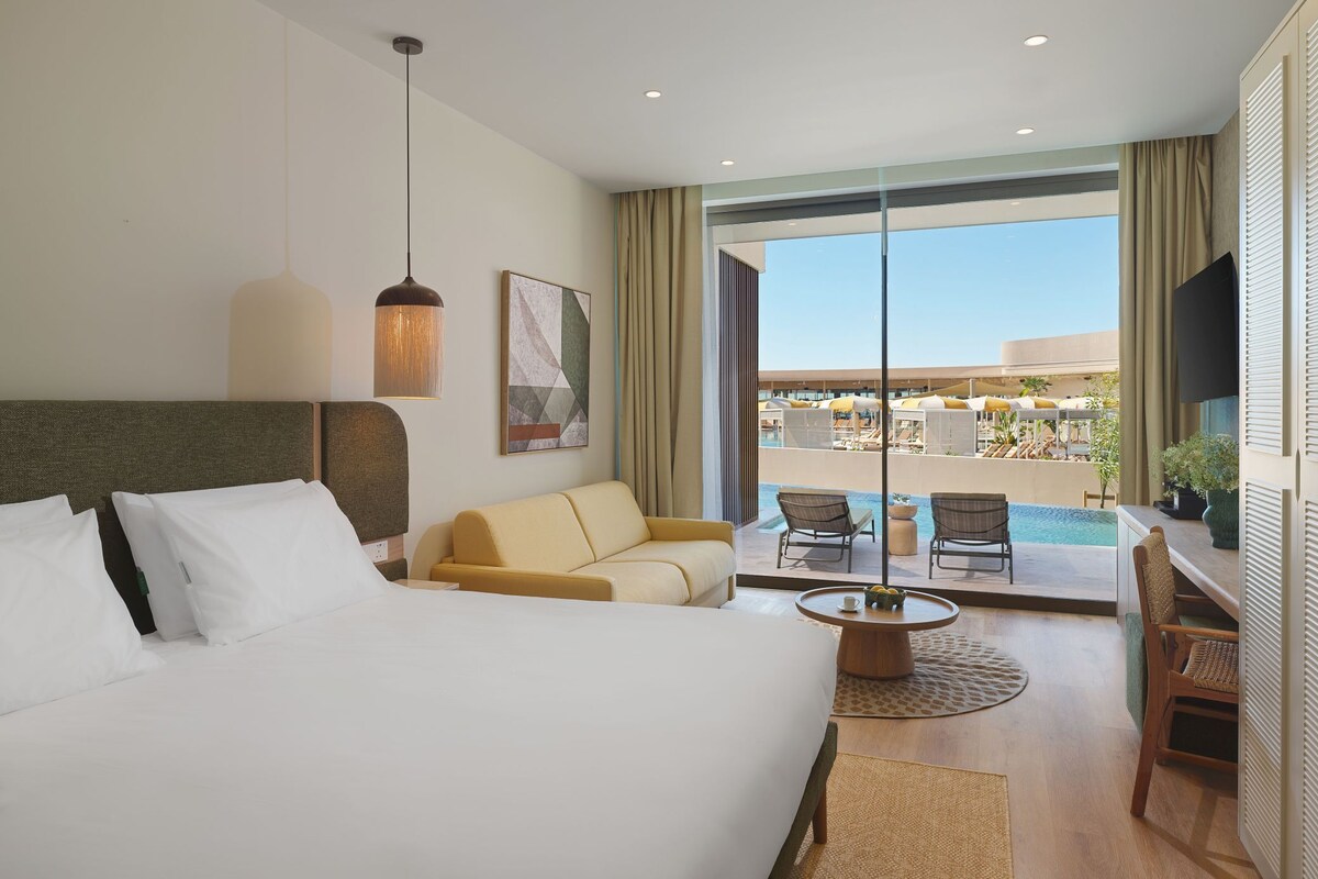 Ein Hotelzimmer mit Bett, Sofa, Esstisch, Fernseher und Blick auf einen Pool und Sitzgelegenheiten im Freien.