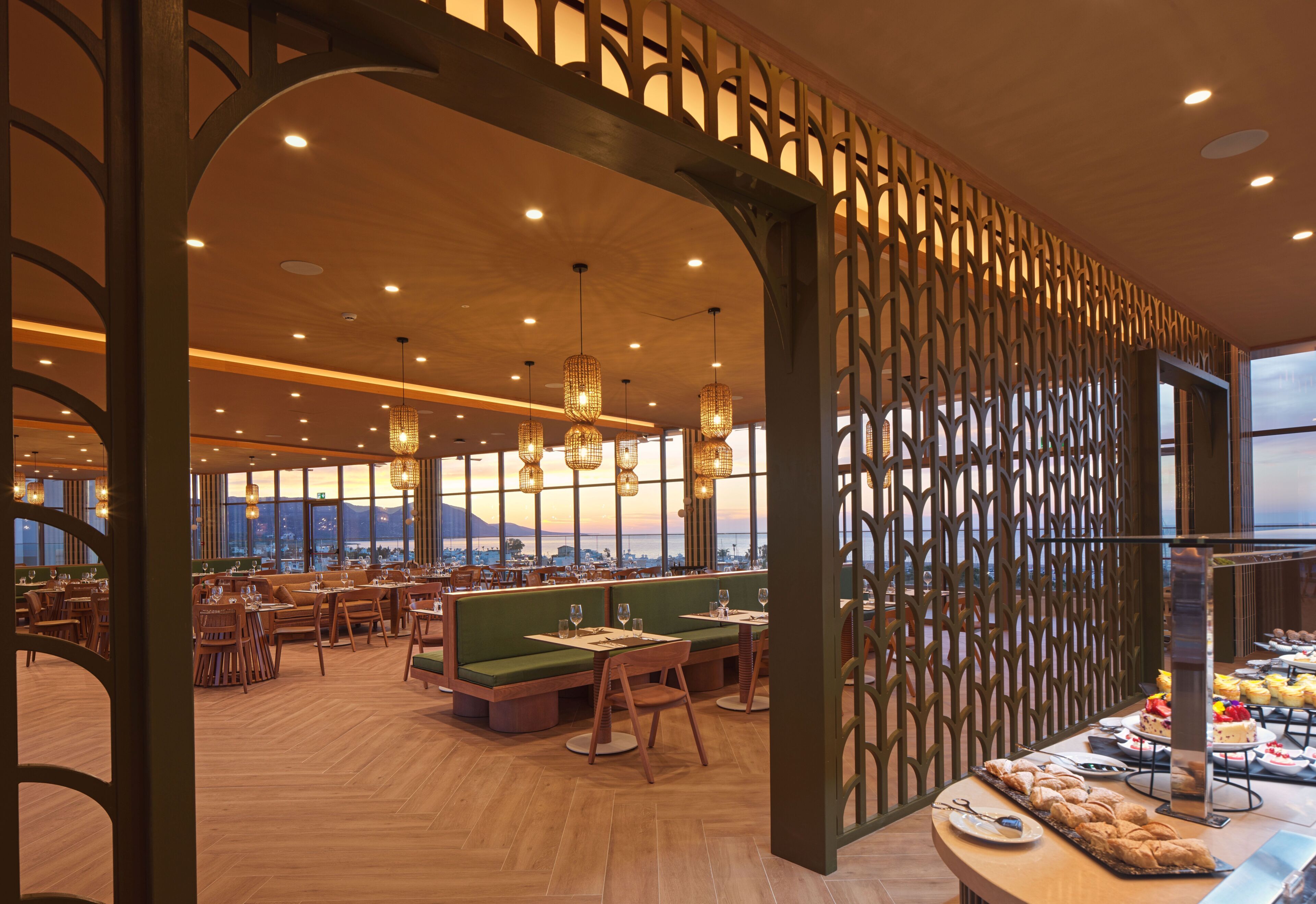 Interior de un restaurante con suelo de madera, asientos verdes y vistas de la ciudad a través de grandes ventanales.