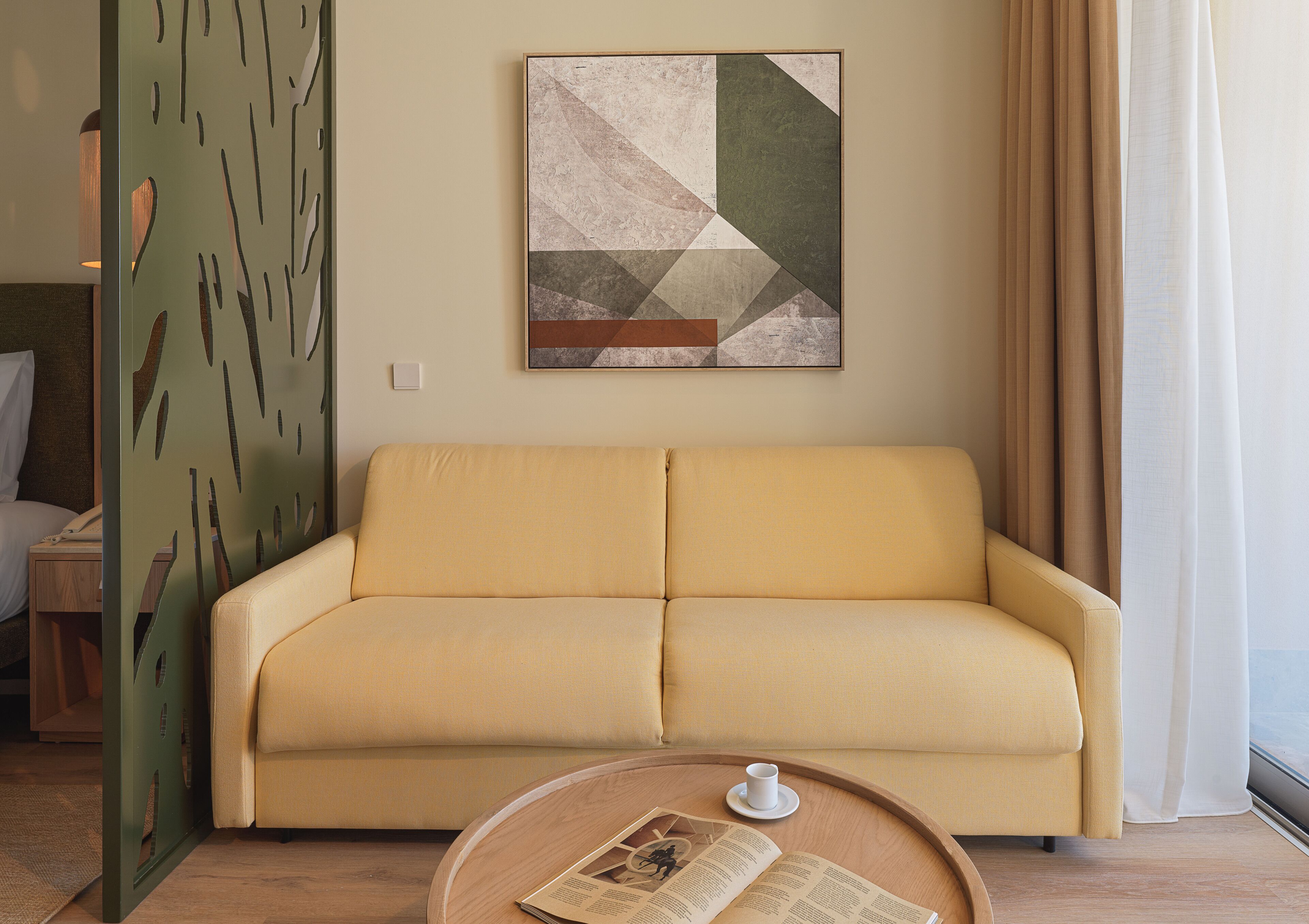 Un sofá color beige en una habitación con un cuadro abstracto geométrico encima, una mesita redonda con una taza y un libro, y una ventana con cortinas.