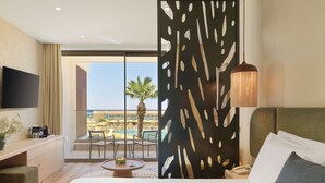 A hotel room with a bed, a desk, a TV, a decorative screen, a lamp, and a view of a pool and palm tree. - NALU Latchi Leonardo Limited Edition (Polis)