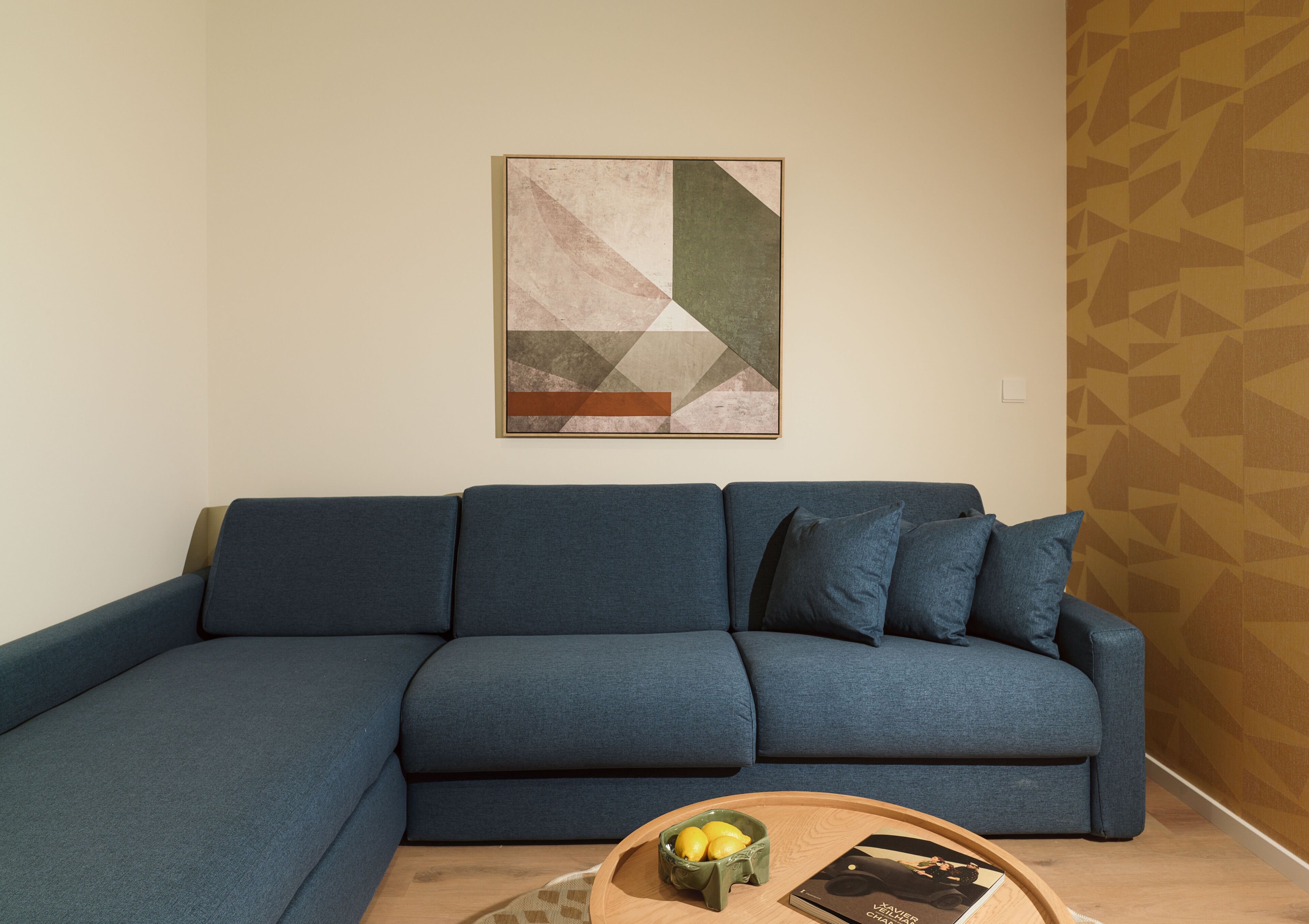 Una sala de estar con un sofá seccional, una mesa de centro con un libro y un cuenco de limones, y un cuadro abstracto geométrico en la pared.