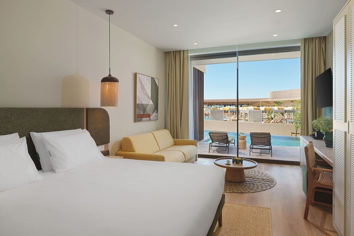 Habitación de hotel con cama, sofá, mesa de comedor, televisión y vistas a una piscina y zona de estar al aire libre.