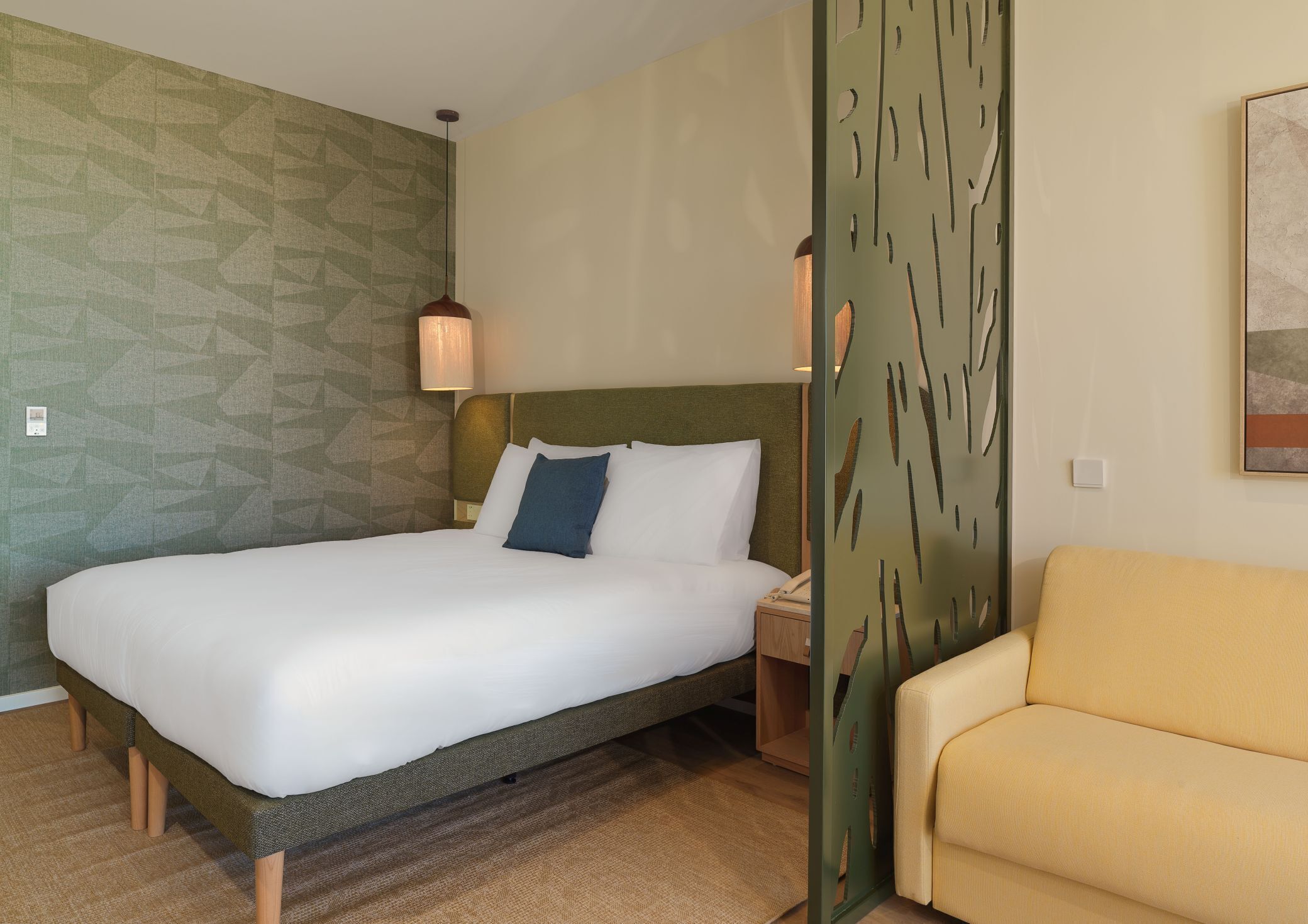 A hotel room with a bed, a nightstand, a lamp, a sofa, and a decorative wall panel.