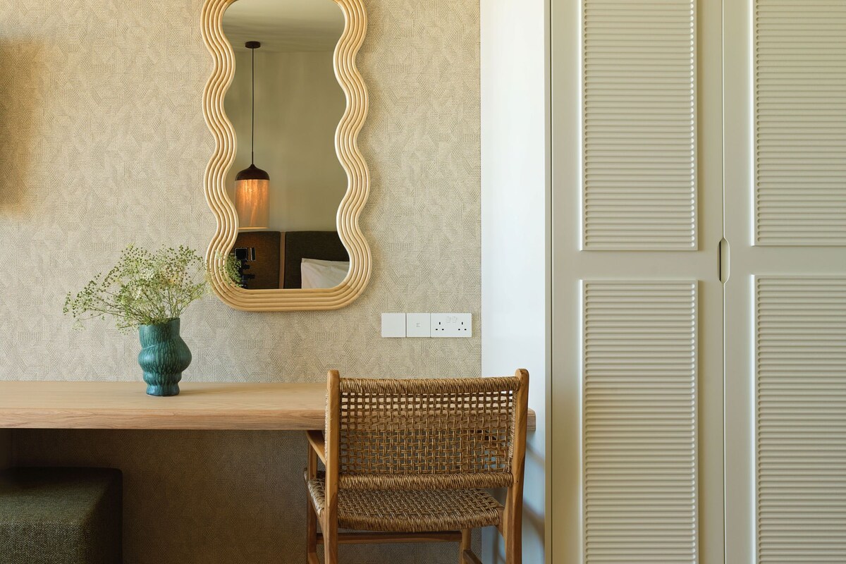A room with a wooden desk, a wicker chair, a mirror, and a hanging light fixture.