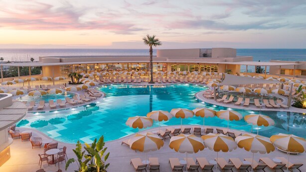 A large indoor pool with multiple sun loungers and umbrellas, surrounded by a modern building with large windows overlooking the sea. - NALU Latchi Leonardo Limited Edition (Polis)