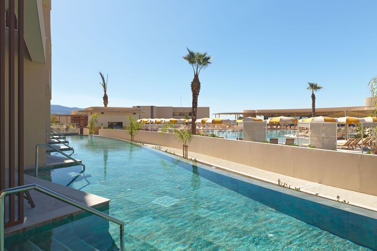 A clear swimming pool with sun loungers and umbrellas, surrounded by palm trees and a clear sky.