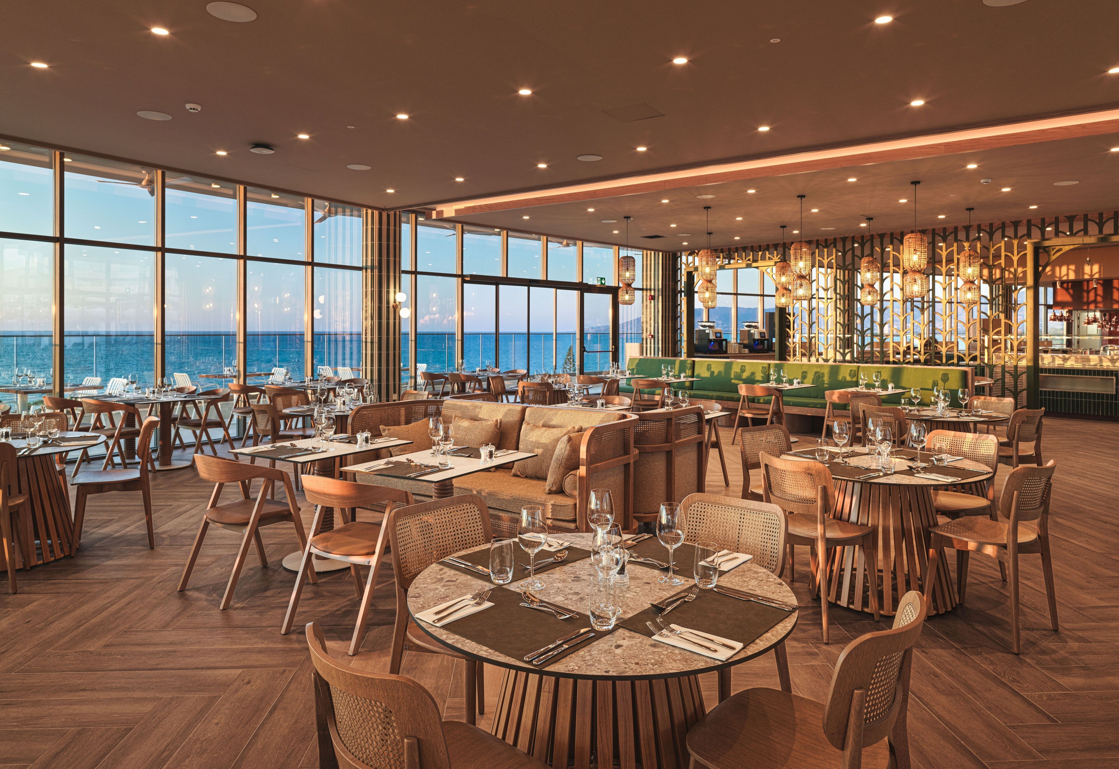 A restaurant with multiple dining areas, large windows overlooking the sea, and a bar area with hanging lights.