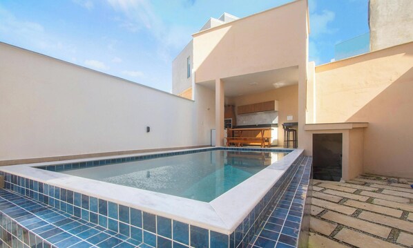 A swimming pool with a tiled edge, a kitchen area, and a patio with stone tiles.