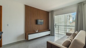 A modern living room with a flat-screen TV mounted on a wooden panel wall, a glass door leading to an outdoor area, and a beige sofa. - Apt in Condo w/ BBQ Area in Porto - BA MAA (Porto Seguro)
