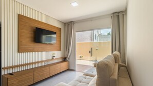 A modern living room with a flat-screen TV mounted on a wooden panel wall, a sofa, and a sliding glass door leading to an outdoor area. - Apt in Condo w/ BBQ Area in Porto - BA MAA (Porto Seguro)