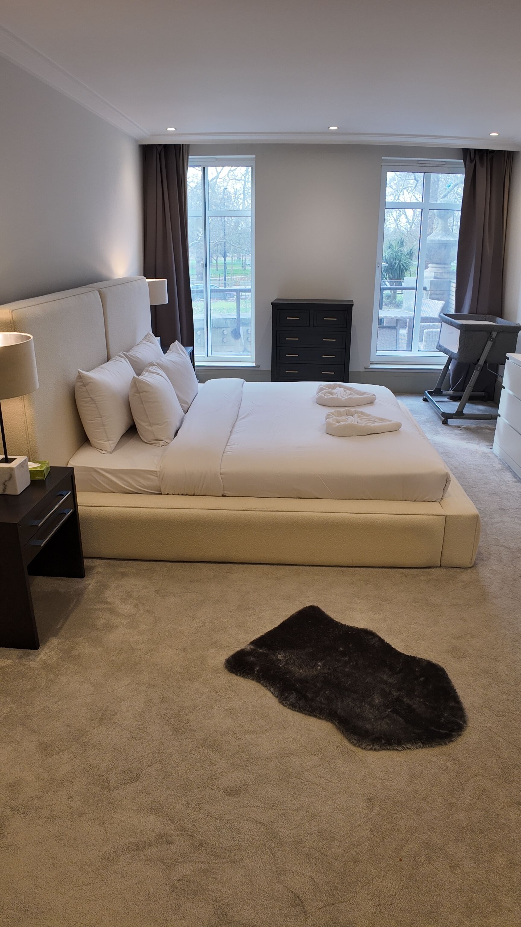 Une chambre à coucher moderne avec un grand lit, une commode sombre et une vue sur l’extérieur grâce à de grandes fenêtres.