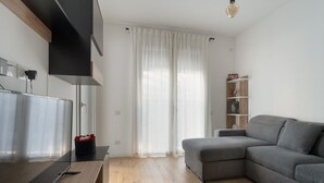 Ein modernes Wohnzimmer mit einem grauen Sofa, Holzböden, einem Fernseher auf einem Holztisch und einem weißen Vorhang.