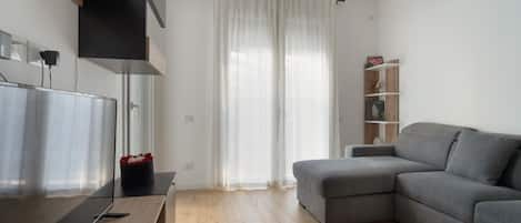 Un salón moderno con un sofá gris, suelo de madera, un televisor sobre un mueble de madera y una cortina blanca.