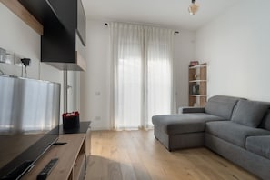 A modern living room with a grey sofa, wooden flooring, a TV on a wooden stand, and a white curtain. - GuestHost - A comfortable and bright 55 square meters apartment, perfect for four people, located on the first floor of a building with an elevator (NOT accessible to disabled guests).The property is located in the Cascina Merlata neighborhood, a growing (Milan)