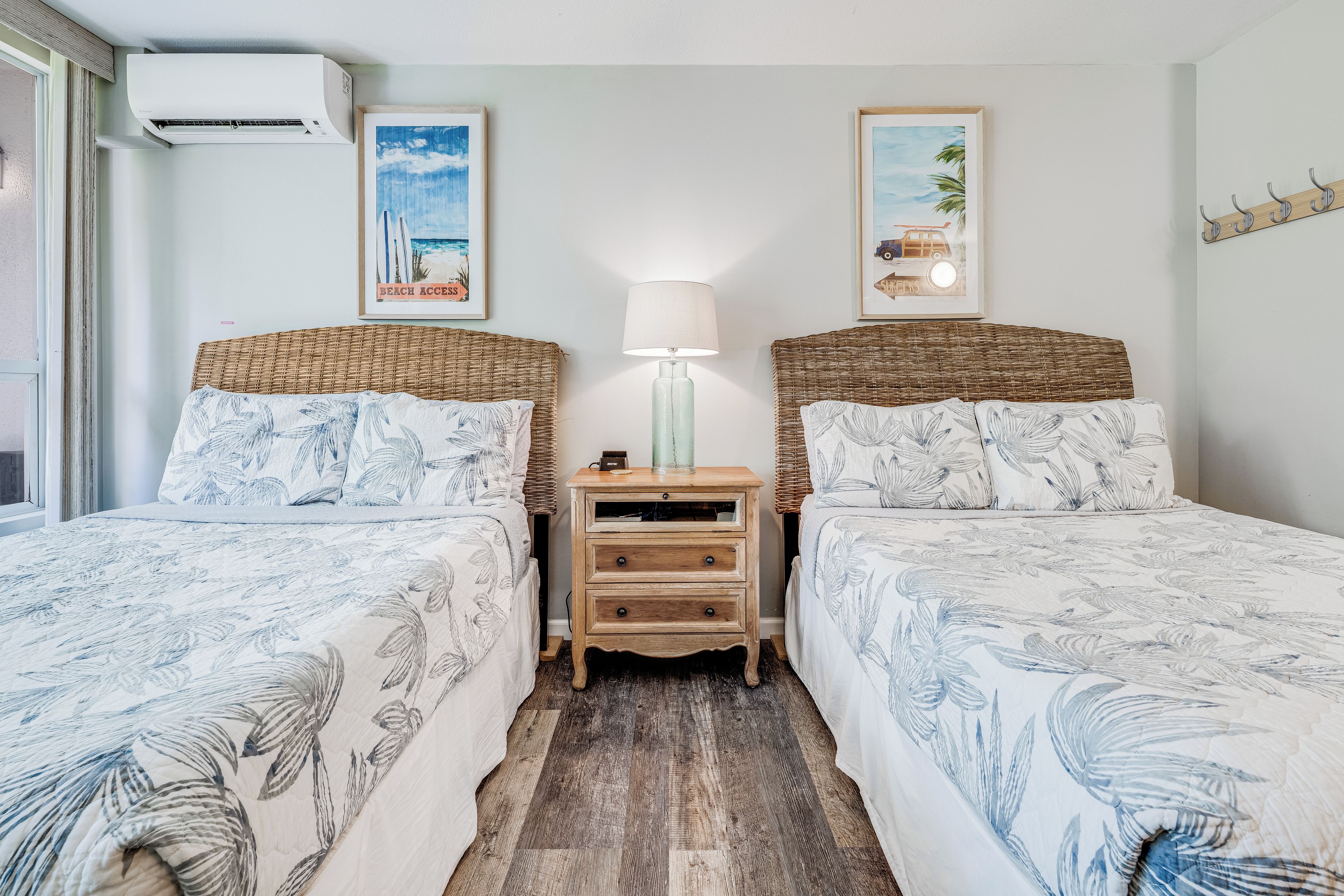 Una habitación con dos camas, una mesita de noche con lámpara y decoración en la pared que representa escenas de playa.