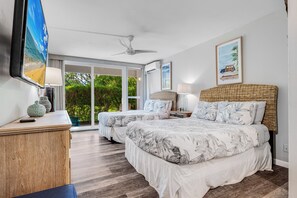 Ein Schlafzimmer mit zwei Betten, einem Fernseher, einem Deckenventilator und einem großen Fenster mit Blick auf Grünflächen.