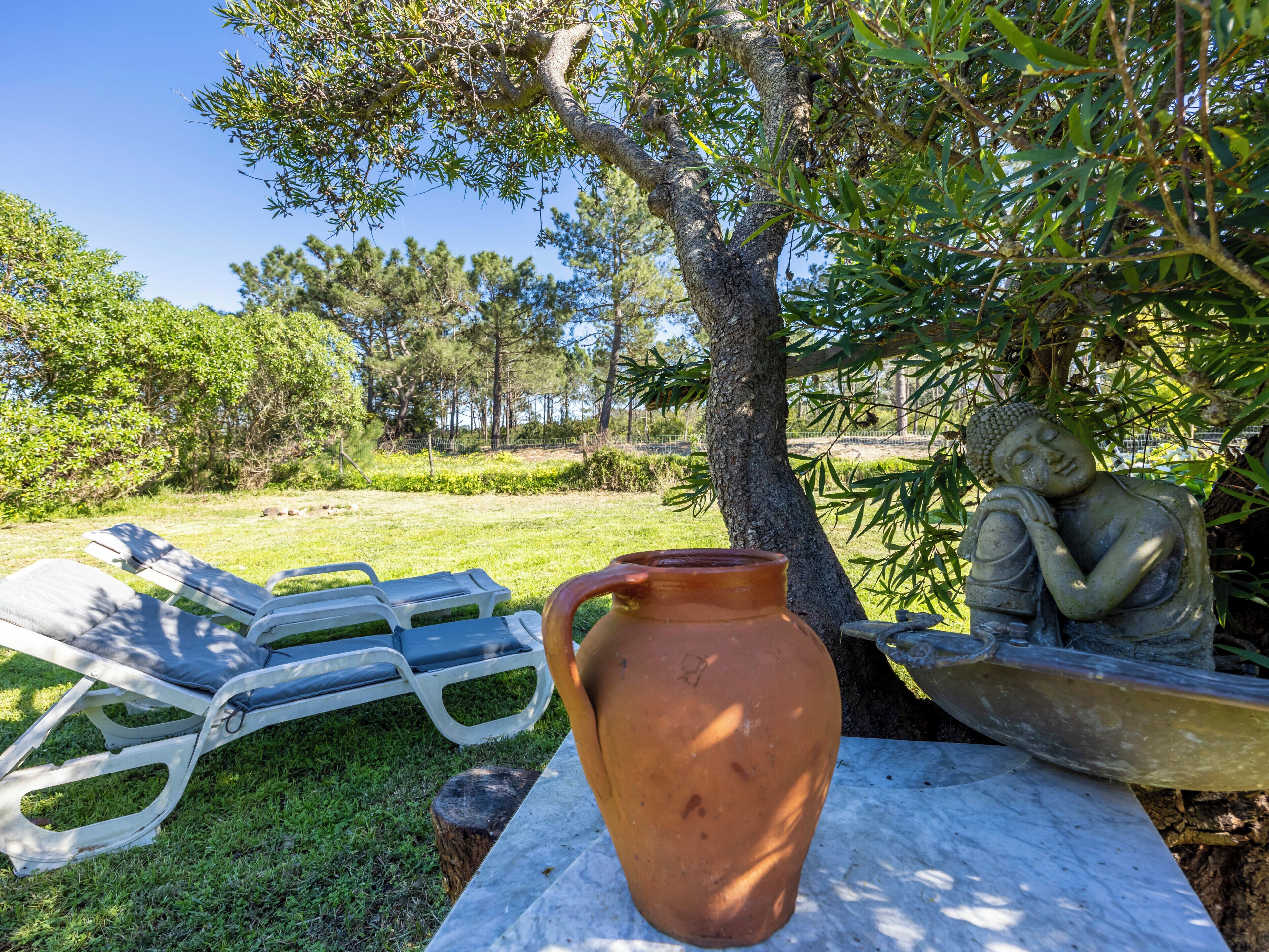 Een terracotta pot op een tafel, een Boeddhabeeld en twee witte lounge fauteuils onder een boom.