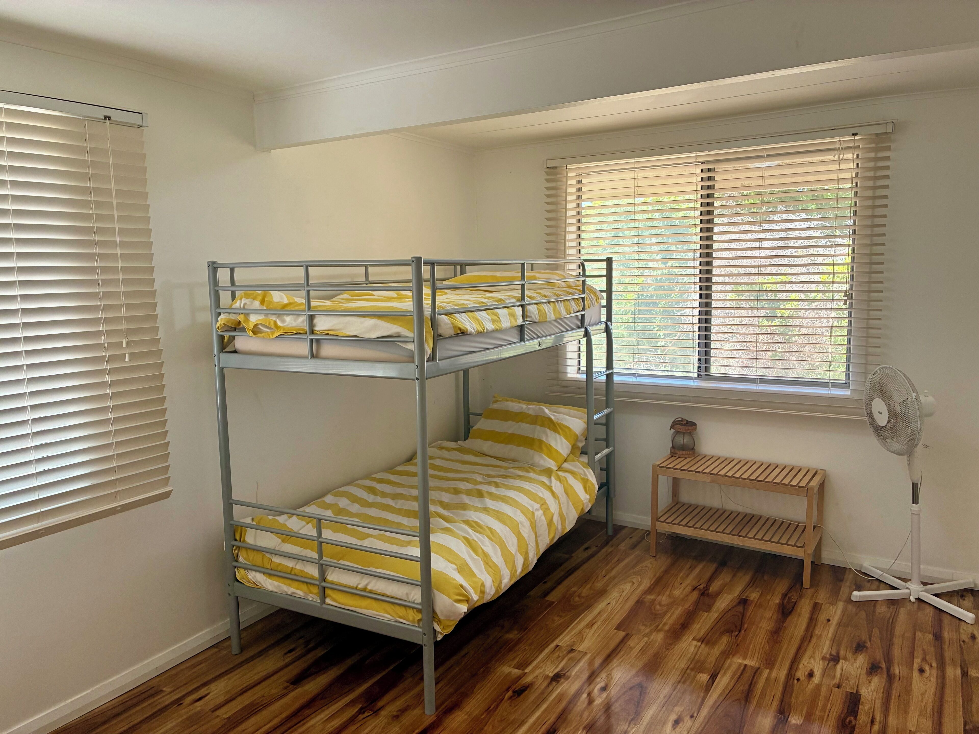 Habitación con literas, suelo de madera, un ventilador de pie y una ventana con persianas.
