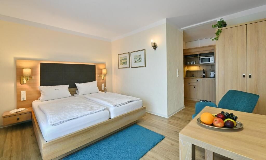 A modern hotel room with a bed, bedside tables, a desk, a chair, a small kitchenette, and a fruit bowl.