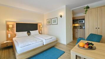A modern hotel room with a bed, bedside tables, a desk, a chair, a small kitchenette, and a fruit bowl.