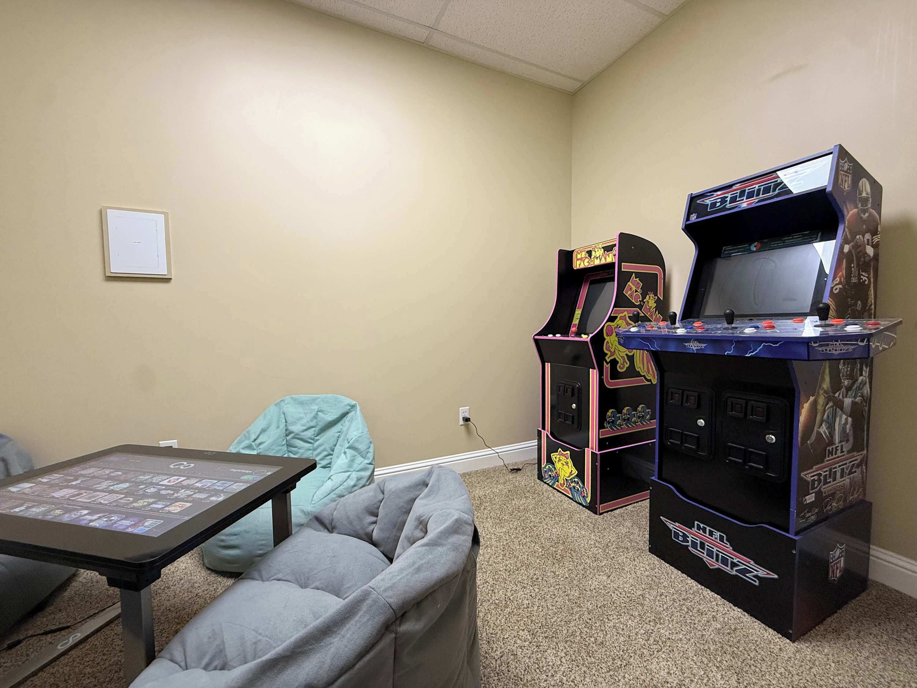 Una habitación con paredes color beige, un sofá gris, una mesita con una pantalla y dos máquinas de videojuegos con la etiqueta 'NFL Blitz'.