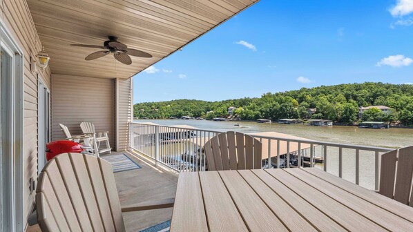 A wooden deck with a table and chairs, overlooking a river with boats and houses in the background.