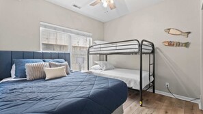 A bedroom with a bunk bed, a large bed with a blue quilt, and a window with a view of the outside.