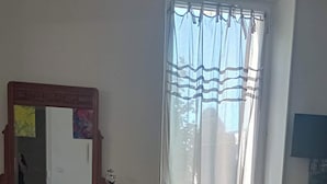 Una habitación con cómoda de madera, cama con colcha floral, ventana con cortinas translúcidas y televisor en la pared.