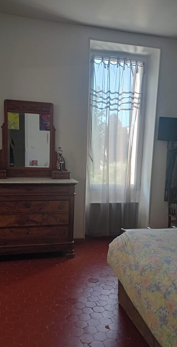A room with a wooden dresser, a bed with a floral bedspread, a window with sheer curtains, and a television on the wall. - A lovely little house with a garden and a pool (Salon-de-Provence)