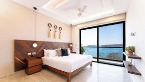 Een moderne slaapkamer met een groot bed, houten hoofdeinde, nachtkastjes, een plafondventilator en een groot raam met uitzicht op zee.