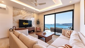 Een moderne woonkamer met een grote flatscreen televisie, een zithoek, een salontafel en uitzicht op de zee door grote ramen.