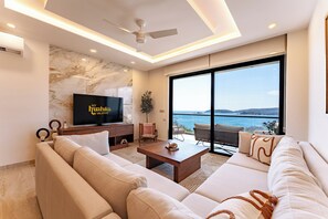 Ein modernes Wohnzimmer mit einem großen Flachbildfernseher, einer Sofagarnitur, einem Couchtisch und Meerblick durch große Fenster.
