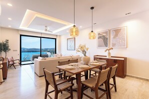 Ein moderner Essbereich mit einem großen Tisch, Stühlen und Pendelleuchten. Ein Sofa und ein großes Fenster bieten Blick ins Freie.