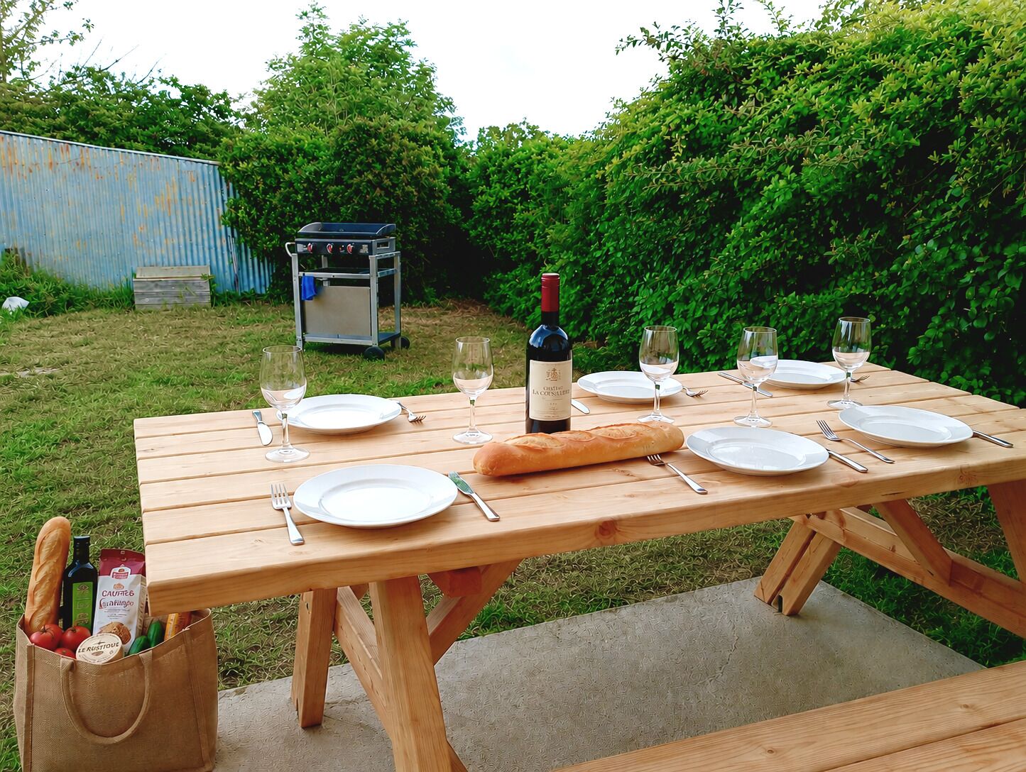 A wooden picnic table set with plates, wine glasses, a baguette, and a bottle of wine.