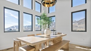 A spacious dining area with a wooden table and benches, large windows offering mountain views, and a modern pendant light. - Luxury Deer Valley East Village Retreat Hot Tub 4 Bedroom Home (Heber City)