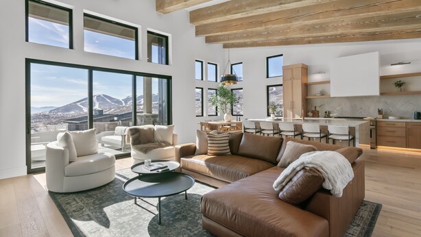 A spacious living area with a large window, a brown leather sofa, a white armchair, and a dining area with a kitchen in the background. - Luxury Deer Valley East Village Retreat Hot Tub 4 Bedroom Home (Heber City)