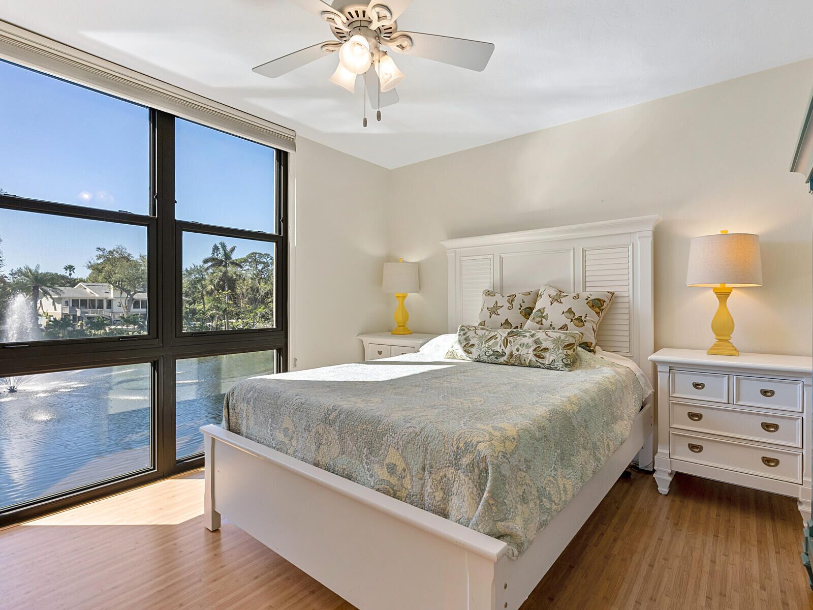 A bedroom with a large window, a bed, a nightstand, a lamp, and a ceiling fan.