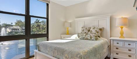 A bedroom with a large window, a bed, a nightstand, a lamp, and a ceiling fan.