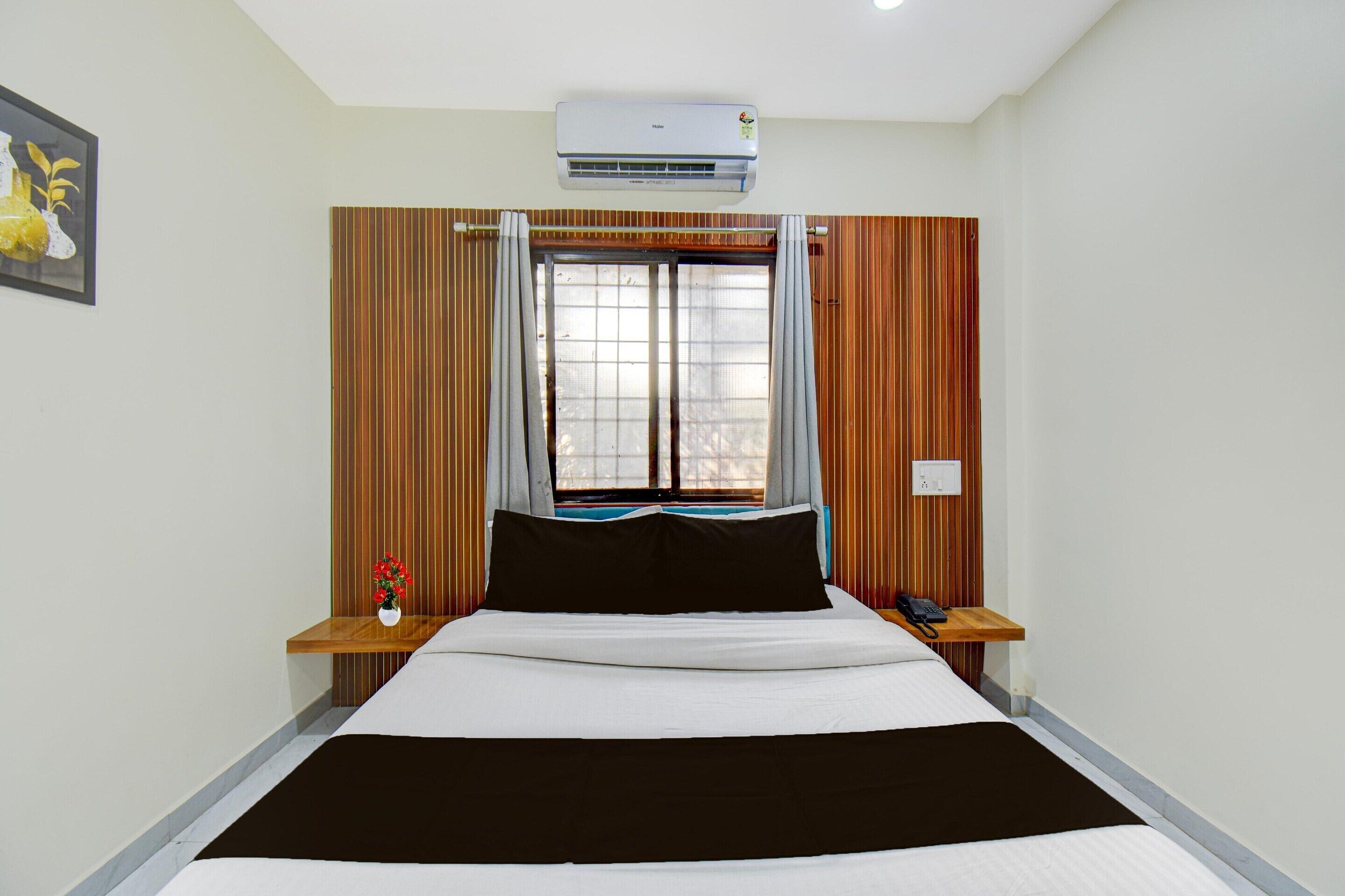 Een hotelkamer met een bed, een airconditioner, een raam met gordijnen, een nachtkastje met telefoon, en een wandcontactdoos.
