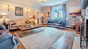A spacious living room with a large sofa, a coffee table, and a fireplace. There are two armchairs and a side table with a lamp. The room has a chandelier and artwork on the walls. - Stunning English country house in rural Kent (Eastry)