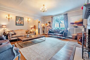 A spacious living room with a large sofa, a coffee table, and a fireplace. There are two armchairs and a side table with a lamp. The room has a chandelier and artwork on the walls. - Stunning English country house in rural Kent (Eastry)