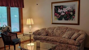 Un salon avec un canapé à motifs floraux, une table basse en verre, une chaise en bois foncé, une lampe et un ventilateur de plafond.
