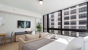 Una habitación de hotel moderna con una cama grande, un escritorio con una silla, televisión y vistas a un edificio a través de ventanas amplias.