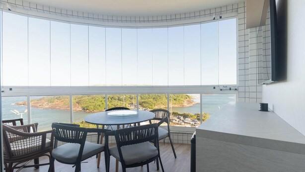 A modern room with a curved ceiling, a round table with chairs, and a large window offering a view of a coastline and water. - Building w/ Pool in Front of the Beaches of Bacutia and Peracanga - Emqs (Guarapari)