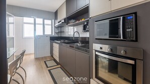 A modern kitchen with a microwave, oven, and stainless steel appliances. There is a window providing natural light and a view of the outside. - Building w/ Pool in Front of the Beaches of Bacutia and Peracanga - Emqs (Guarapari)
