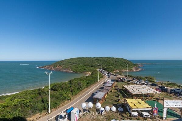 An aerial view of a coastal area with a road leading to a beach, parked vehicles, and a signboard. - Building w/ Pool in Front of the Beaches of Bacutia and Peracanga - Emqs (Guarapari)
