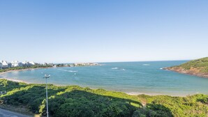 A coastal view with a beach, clear blue water, and a cityscape in the background. - Building w/ Pool in Front of the Beaches of Bacutia and Peracanga - Emqs (Guarapari)