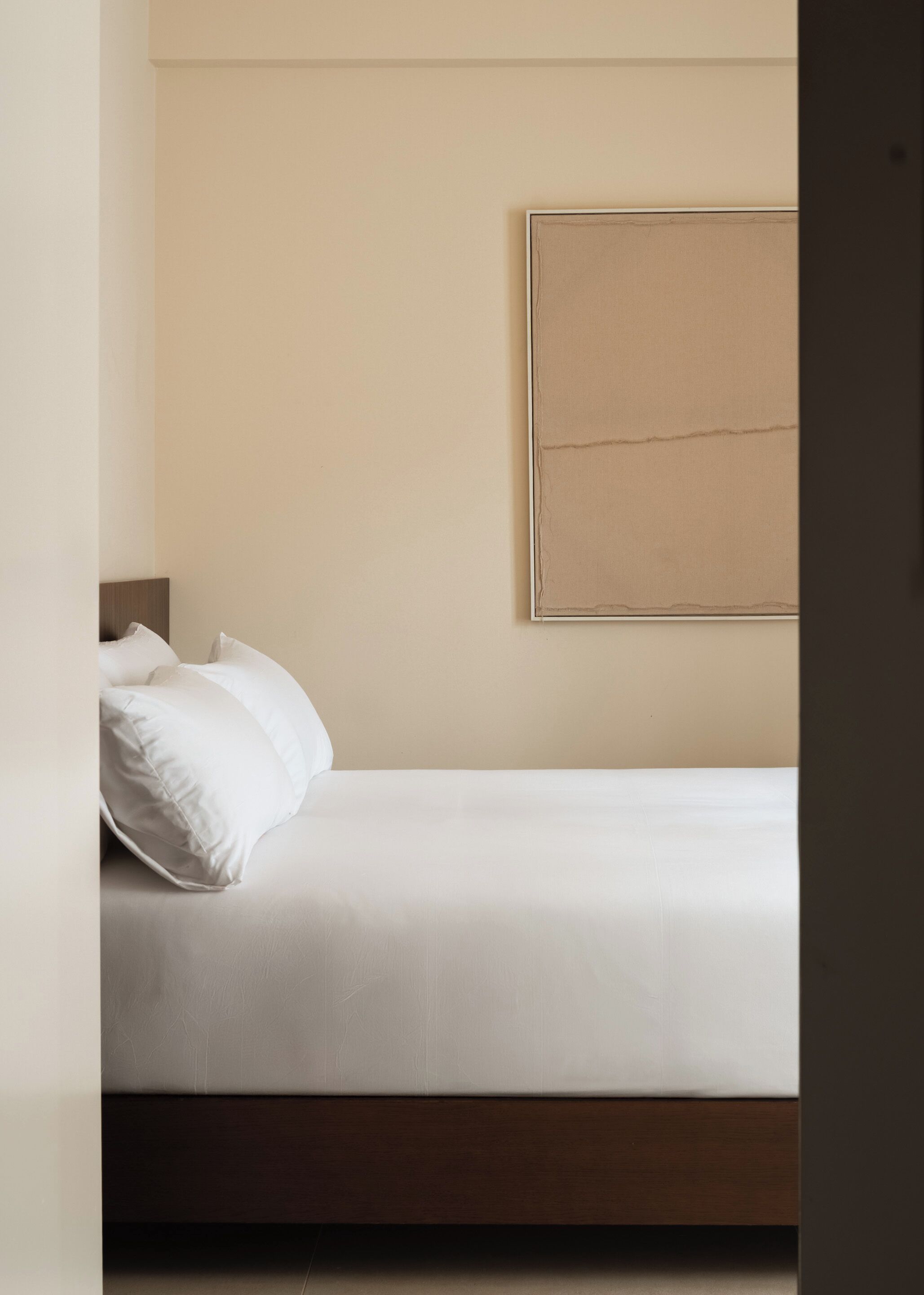Un lit simple avec une literie et des oreillers blancs, une tête de lit en bois et un mur de couleur beige sobre orné d’une œuvre d’art encadrée.