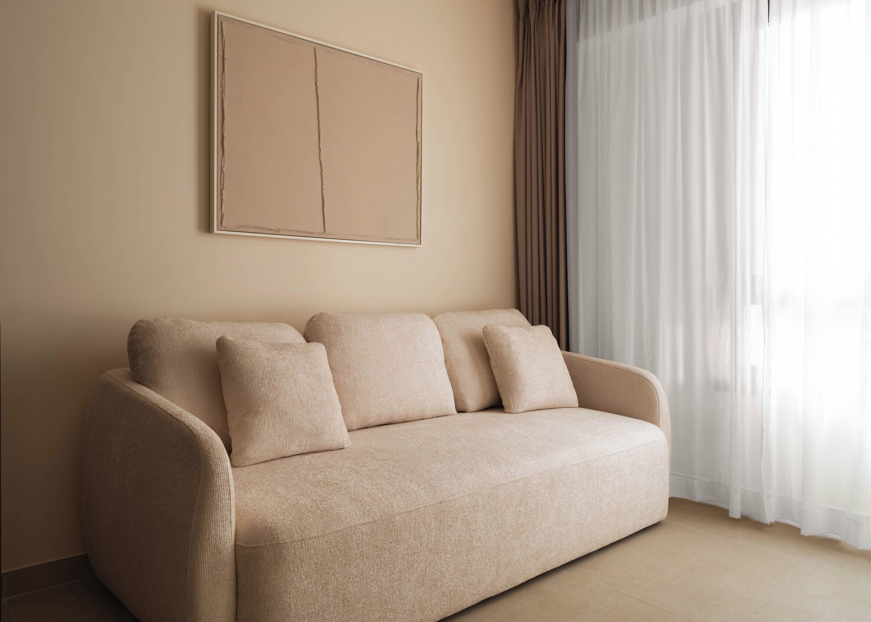 A beige sofa with four cushions in a room with a plain wall and a window with sheer curtains.