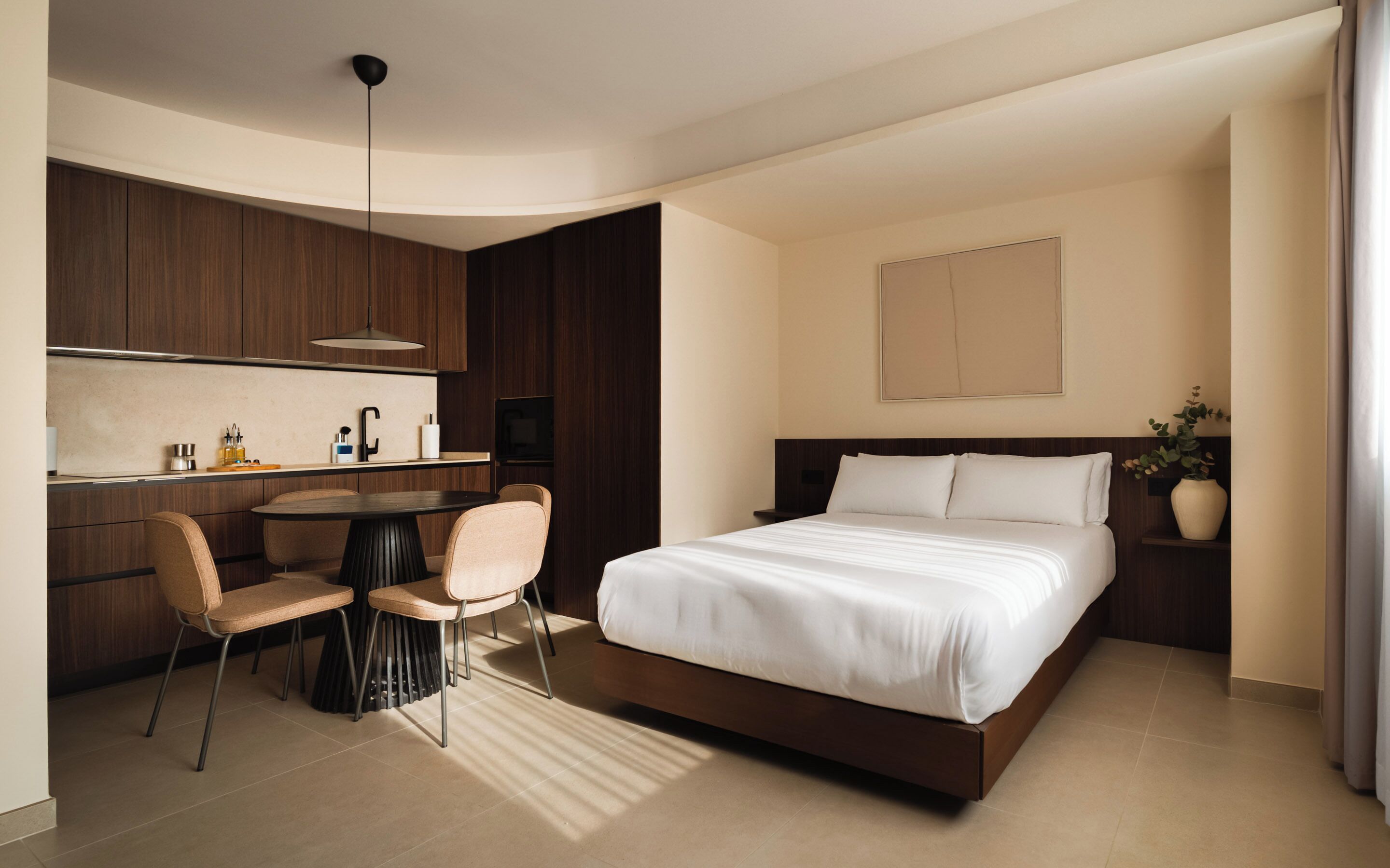 A modern hotel room with a bed, a desk, chairs, a pendant light, and a potted plant.